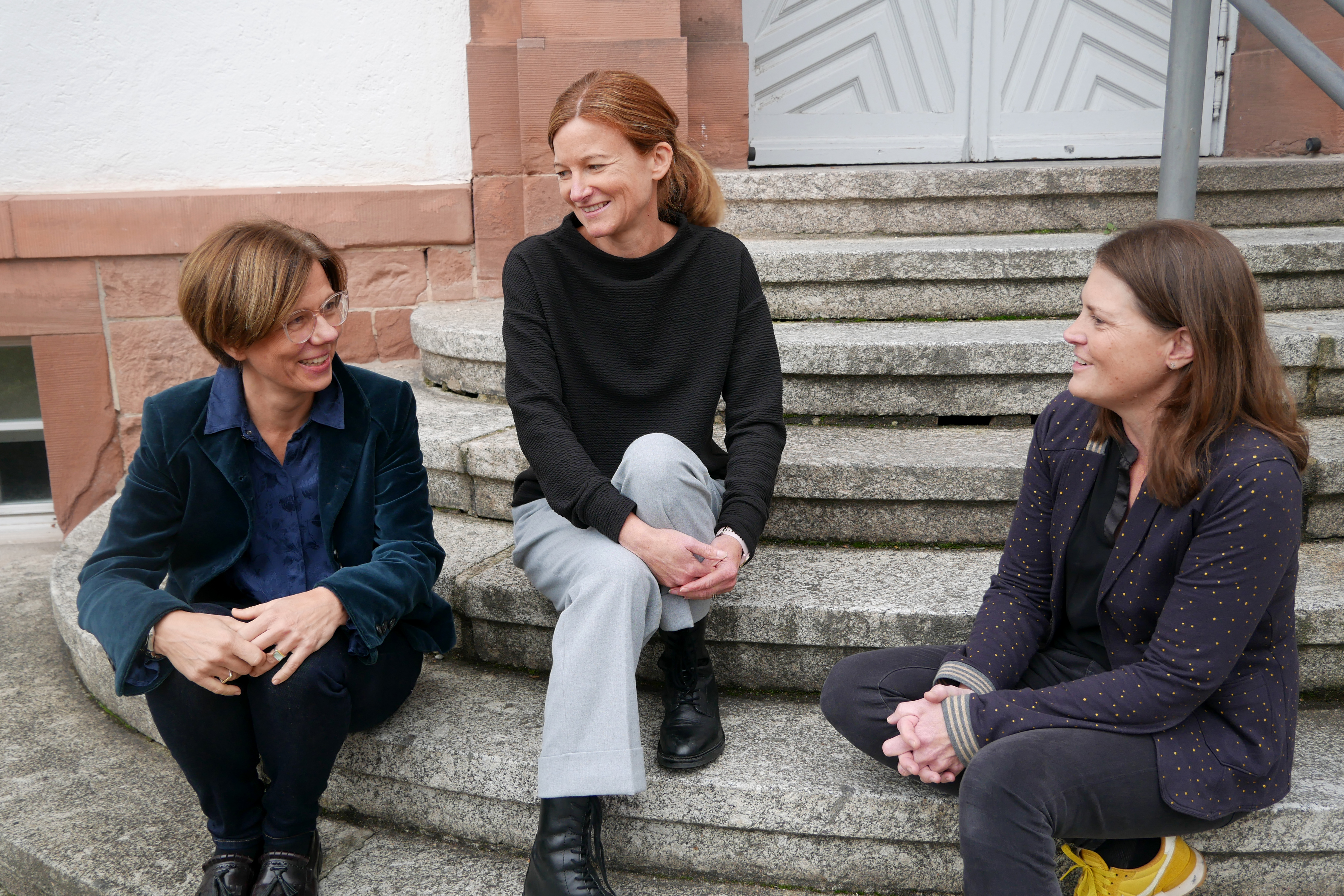 Fachrichtung LBS Die drei Ausbilderinnen der Fachrichtung Lernen bei Blindheit und Sehbehinderung sitzen auf einer Steintreppe am Eingang des Seminargebäudes und unterhalten sich.